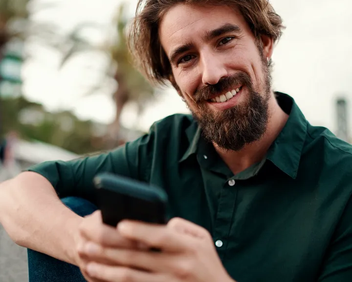 Ein lächelnder Mann mit Bart sitzt im Freien und hält ein Smartphone in der Hand.