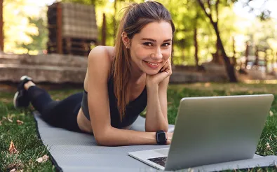Eine junge Frau in Sportkleidung liegt auf einer Gymnastikmatte im Park vor einem aufgeklappten Laptop.