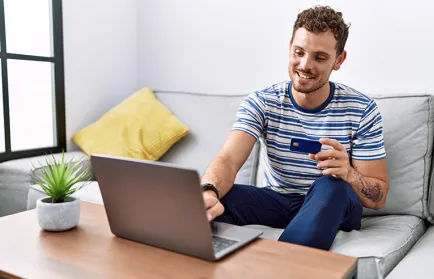 Ein Mann sitzt mit einer Bankkarte in der Hand auf dem Sofa vor einem Laptop auf dem Wohnzimmertisch.