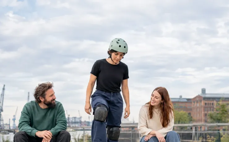 Eine Frau steht mit dem Skateboard an der Kante einer Betonschale, zwei Freunde sehen ihr ermutigend zu