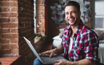 Ein Mann mit Karohemd sitzt lachend mit einem Laptop auf den Knien vor einer Backsteinwand.