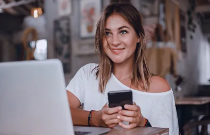 Eine Frau sitzt lächelnd mit einem Smartphone in der Hand vor einem Laptop in einem Café.