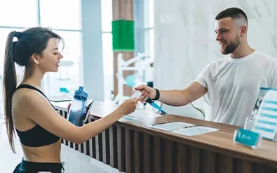 Empfang im Sportstudio: Ein Mitarbeiter reicht einer Kundin am Tresen ihre Unterlagen