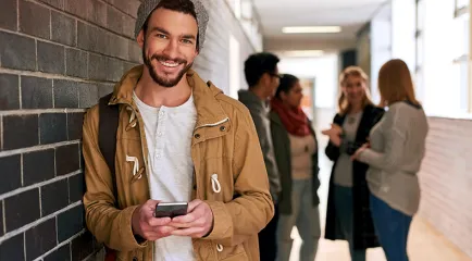 Ein junger Mann mit Mütze lehnt mit einem Smartphone in der Hand an einer Backsteinwand; im Hintergrund unterhalten sich vier Personen.