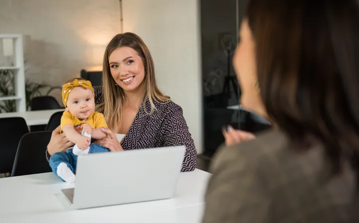 Eine junge Mutter hält ihr Baby auf dem Schoß und lächelt vor einem Laptop einer Gesprächspartnerin zu.