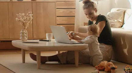 Eine Frau sitzt mit ihrem Kleinkind auf dem Boden und arbeitet am Laptop auf dem Couchtisch.