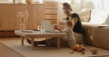 Eine Mutter arbeitet zu Hause am Laptop, während ihr Kleinkind neben ihr am Couchtisch spielt.