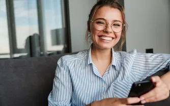 Eine lächelnde Frau mit Brille sitzt auf einem Sofa und hält ein Smartphone in der Hand.