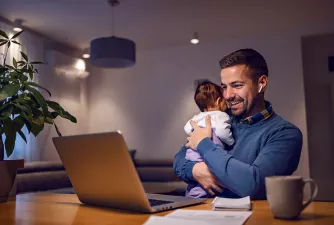 Studium pausieren Ein Mann sitzt mit Baby im Arm vor einem Laptop