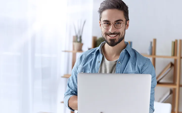 Ein Mann mit Brille sitzt in einem hellen Wohnraum hinter einem aufgeklappten Laptop und schaut lächelnd in die Kamera.