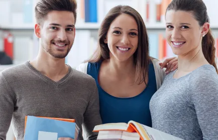 Drei Studierende schauen lachend in die Kamera & halten ihre Bücher in der Hand