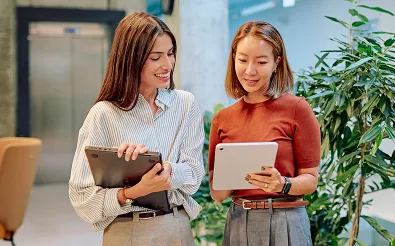 Zwei Frauen stehen lächelnd in einem Büro mit Pflanzen und schauen gemeinsam auf ein Tablet und einen Laptop.