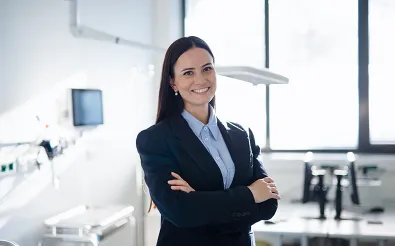 Eine Frau im Blazer steht mit verschränkten Armen in einem hellen Klinikraum und schaut in die Kamera; im Hintergrund medizinische Geräte.