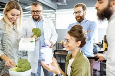 Vier Fachleute betrachten in einem Büro Brokkoli und Nahrungsergänzungsdosen und besprechen deren Zusammensetzung.