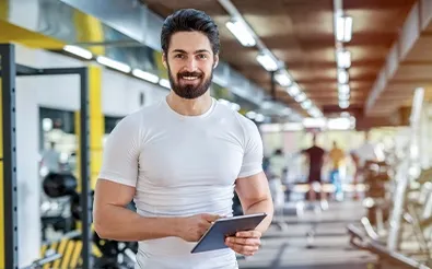 Ein Fitnesstrainer in weißem Shirt hält in einem Fitnessstudio ein Tablet und lächelt in die Kamera.