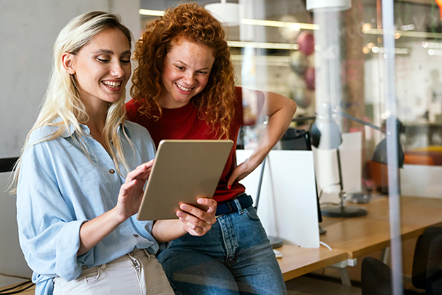 Zwei Frauen stehen nebeneinander in einem Büro und schauen gemeinsam lächelnd auf ein Tablet.