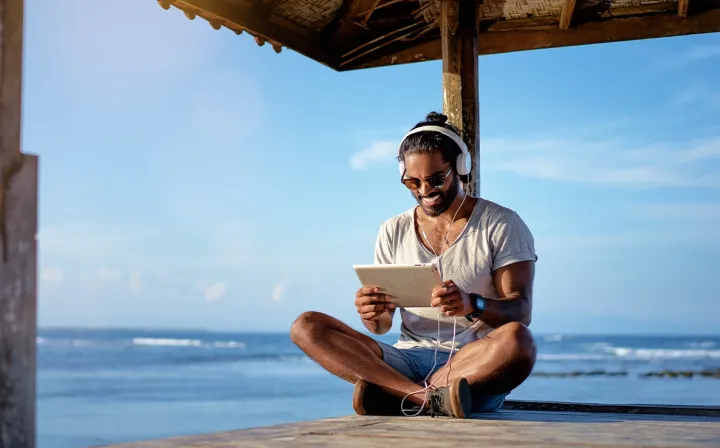 Ein Mann mit Kopfhörern sitzt an einem Holzsteg am Meer und hält ein Tablet in den Händen.