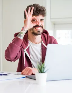 Ein Mann mit Tattoo am Arm hält sich lächelnd die Okay-Geste vors Auge und sitzt dabei an einem Laptop.