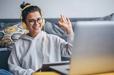 Eine Frau mit Kopfhörern winkt lächelnd in einen Laptop, vor sich ein Notizbuch, im Wohnraum auf dem Sofa.