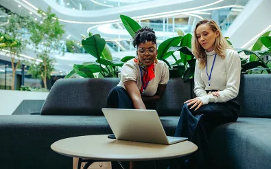 Zwei Frauen sitzen auf einer Sitzbank in einem begrünten Gebäude und schauen gemeinsam auf einen Laptop.