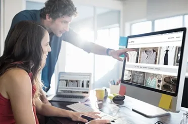 Zwei Personen arbeiten gemeinsam an einem Bildschirm mit einer Online-Shop-Seite; einer zeigt auf ein Produktbild.