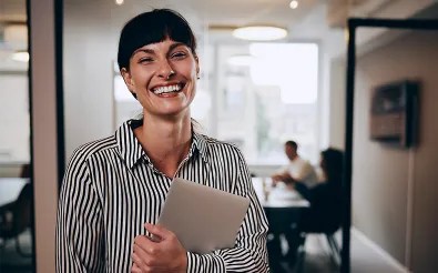 Eine Frau im gestreiften Hemd hält lächelnd ein Tablet im Arm und steht in einem Büro, hinter ihr sind Kollegen am Besprechungstisch zu sehen.