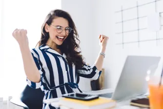 Eine lächelnde Frau mit Brille jubelt mit erhobenen Fäusten am Schreibtisch vor ihrem Laptop.