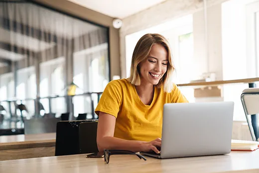 Eine Frau im gelben Shirt sitzt an einem Tisch und tippt lächelnd an ihrem Laptop.