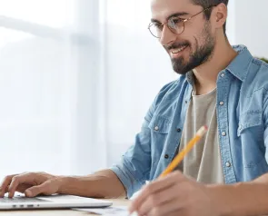 Ein lächelnder Mann mit Brille arbeitet am Laptop und macht sich mit einem Bleistift Notizen.