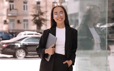 Eine junge Frau im Nadelstreifenanzug steht mit einem Laptop unter dem Arm vor einer Glasfassade und schaut in die Kamera.