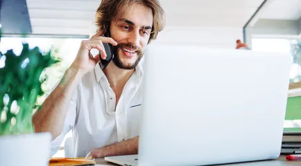 Ein Mann mit Bart telefoniert lächelnd vor seinem Laptop an einem hellen Arbeitsplatz mit Zimmerpflanzen.