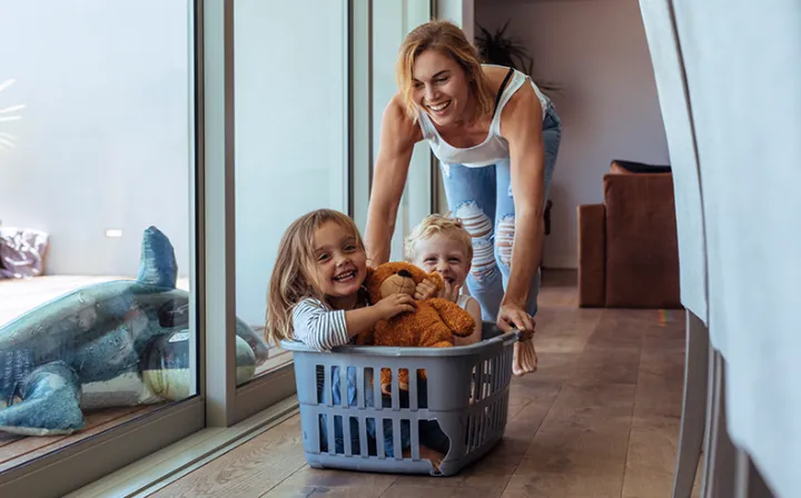 Eine Frau schiebt lachend zwei Kinder in einem Wäschekorb durch die Wohnung.