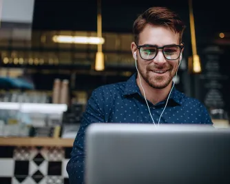 Ein Mann mit Kopfhörern und Brille sitzt lächelnd in einem Café vor einem Laptop und lernt online.