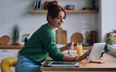 Eine Frau sitzt in einer Küche am Tresen, isst aus einer Schale und schaut dabei auf ein Tablet; daneben ein Glas Orangensaft.