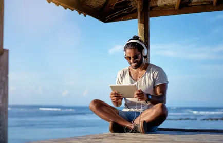 Ein Mann mit Kopfhörern sitzt an einem Holzsteg am Meer und hält ein Tablet in den Händen.