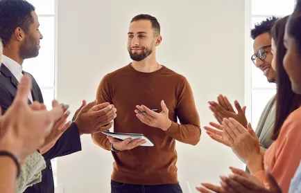 Ein Mann mit Tablet steht lächelnd in einer Gruppe, die ihm applaudiert.