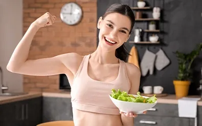 Eine lächelnde junge Frau spannt ihren Oberarm an und hält eine Schüssel mit grünem Salat.