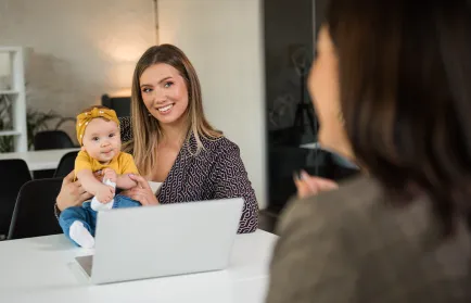 Eine Frau hält ihr Baby auf dem Schoß und lächelt ihrem Gegenüber am Besprechungstisch zu; zwischen ihnen ein aufgeklappter Laptop.
