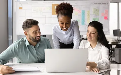Drei Personen arbeiten lächelnd gemeinsam an einem Laptop, eine Frau beugt sich von hinten dazu; im Hintergrund hängt ein Board mit Haftnotizen.