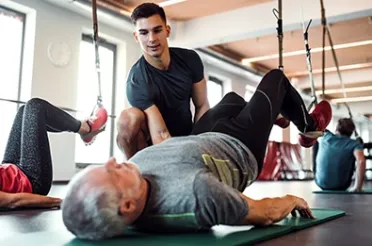 Ein Trainer unterstützt einen älteren Mann bei einer Übung an Schlingen auf einer Trainingsmatte.