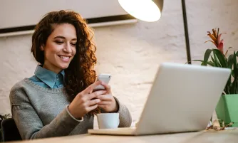 Eine junge Frau mit lockigem Haar sitzt lächelnd am Schreibtisch und schaut auf ihr Smartphone; vor ihr stehen ein Laptop und eine Kaffeetasse.