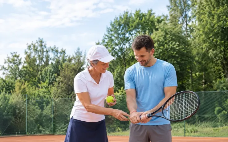 Studien-Tipps der EHiP: Eine Trainerin zeigt einem erwachsenen Anfänger auf dem Sandplatz, wie er den Tennisschläger greift