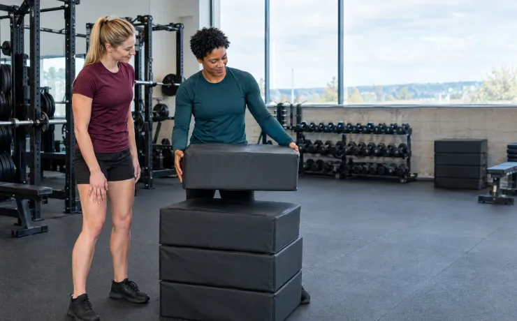 Athletiktrainerin nimmt im Kraftraum eine Sprungbox vom Stapel, nachdem die Athletin schwere Beine gemeldet hat