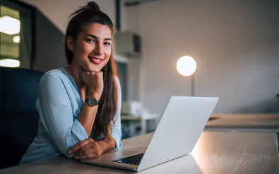 Eine junge Frau sitzt abends am Schreibtisch vor einem Laptop, stützt das Kinn in die Hand und schaut in die Kamera.