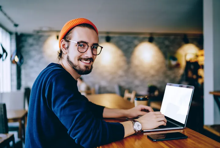 Ein junger Mann mit Mütze und Brille arbeitet in einem Café am Laptop und schaut lächelnd in die Kamera.