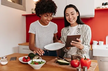 Zwei Frauen stehen in einer Küche, schauen lächelnd gemeinsam auf ein Tablet und bereiten dabei frisches Gemüse zu.