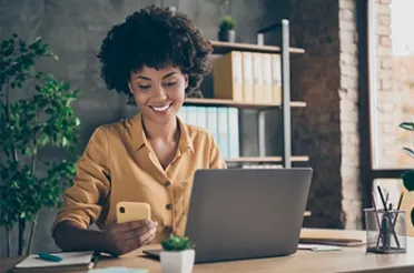 Eine lächelnde Frau sitzt mit Smartphone in der Hand an einem Laptop in einem Büro mit Regal und Pflanzen.