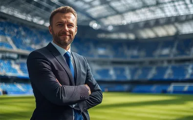 Ein Mann im Anzug steht mit verschränkten Armen am Spielfeldrand eines leeren Fußballstadions.
