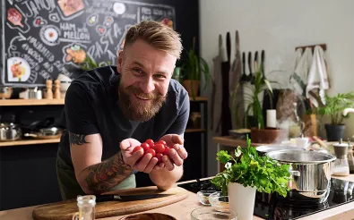 Lächelnder Koch zeigt eine Handvoll Cherrytomaten in einer Küche mit frischen Kräutern
