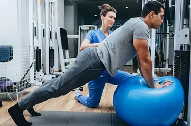 Ein Therapeut stützt sich in einer Physiotherapiepraxis auf einen Gymnastikball, während eine Kollegin die Übung begleitet.